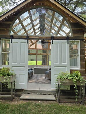 Une structure en bois surmontée d’un grand toit en verre en forme d’arche, avec un banc et des plantes en pot.