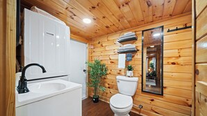 A wooden-paneled bathroom with a white sink, black faucet, white toilet, and a mirror above the toilet. - Hot Tub! Tiny House Retreat in the Woods (Stanton)