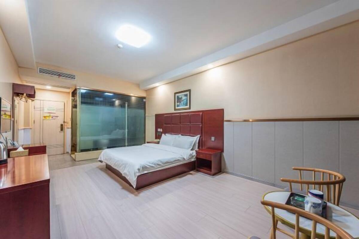 Quarto de hotel com cama, mesa de cabeceira, cadeira de madeira e box de vidro.