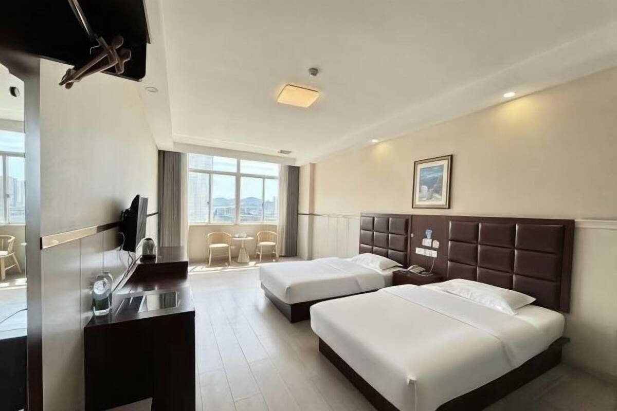 Quarto de hotel com duas camas, uma escrivaninha, uma cadeira, uma janela com vista para a cidade e um quadro na parede.