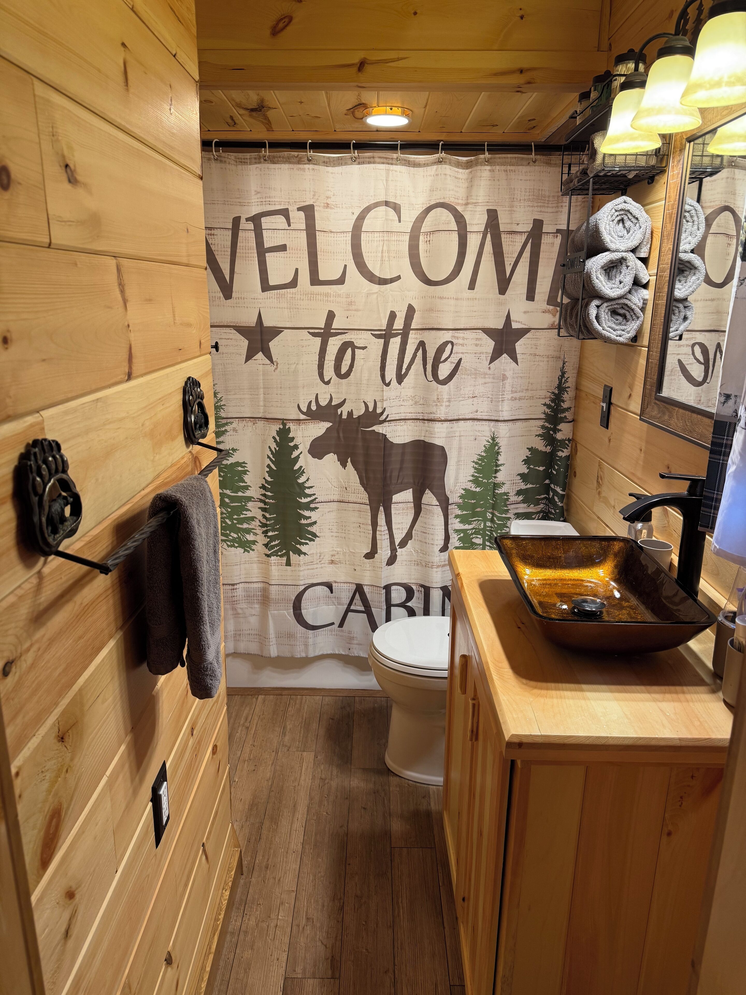 A cabin-style bathroom with a wooden interior, a moose-themed welcome sign, a sink with a glass basin, and towels hanging on hooks.