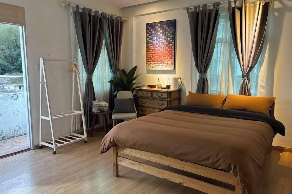 A bedroom with a wooden bed, a bedside table, a chair, a plant, and a window with curtains.