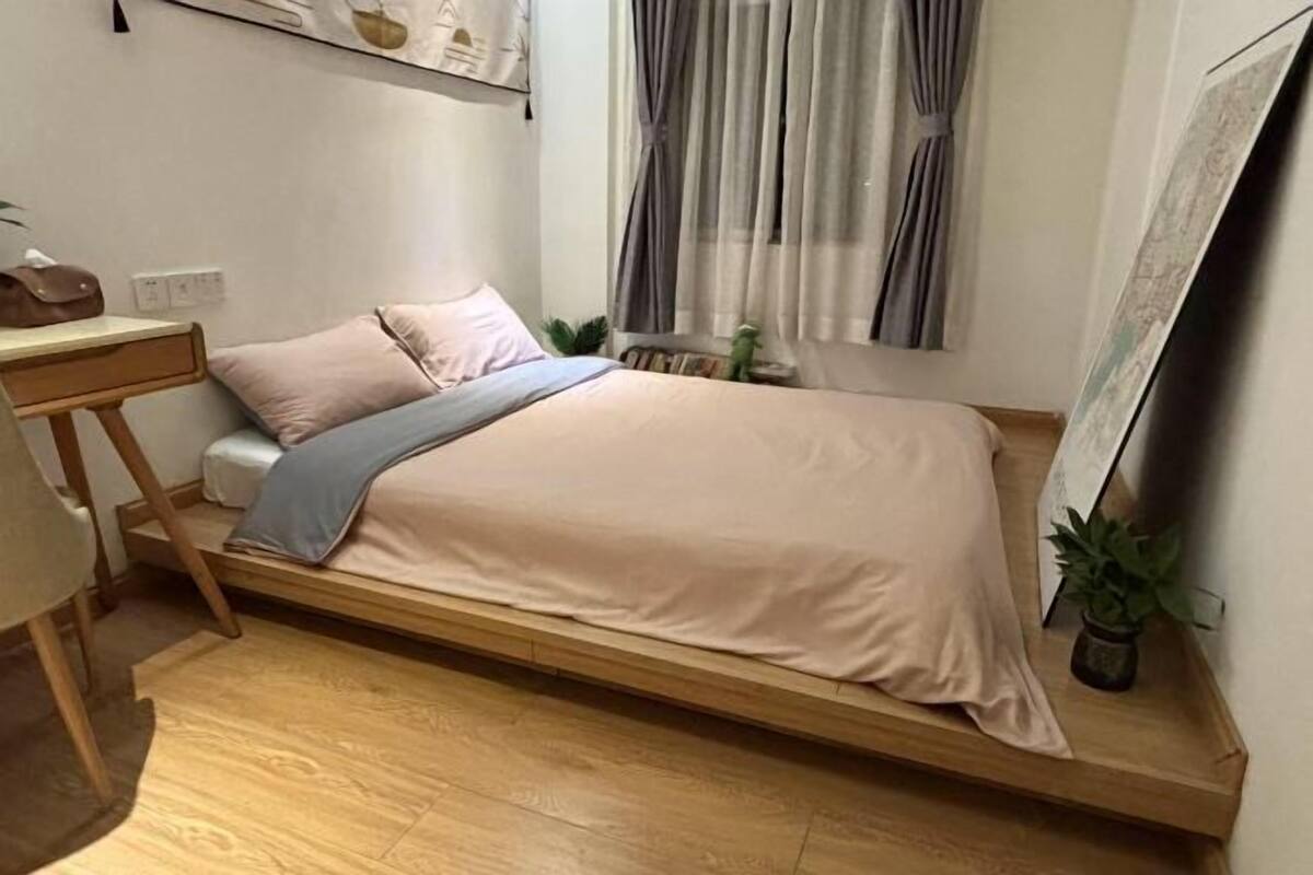 A single bed with a wooden frame, a bedside table, a small plant, and a window with curtains.