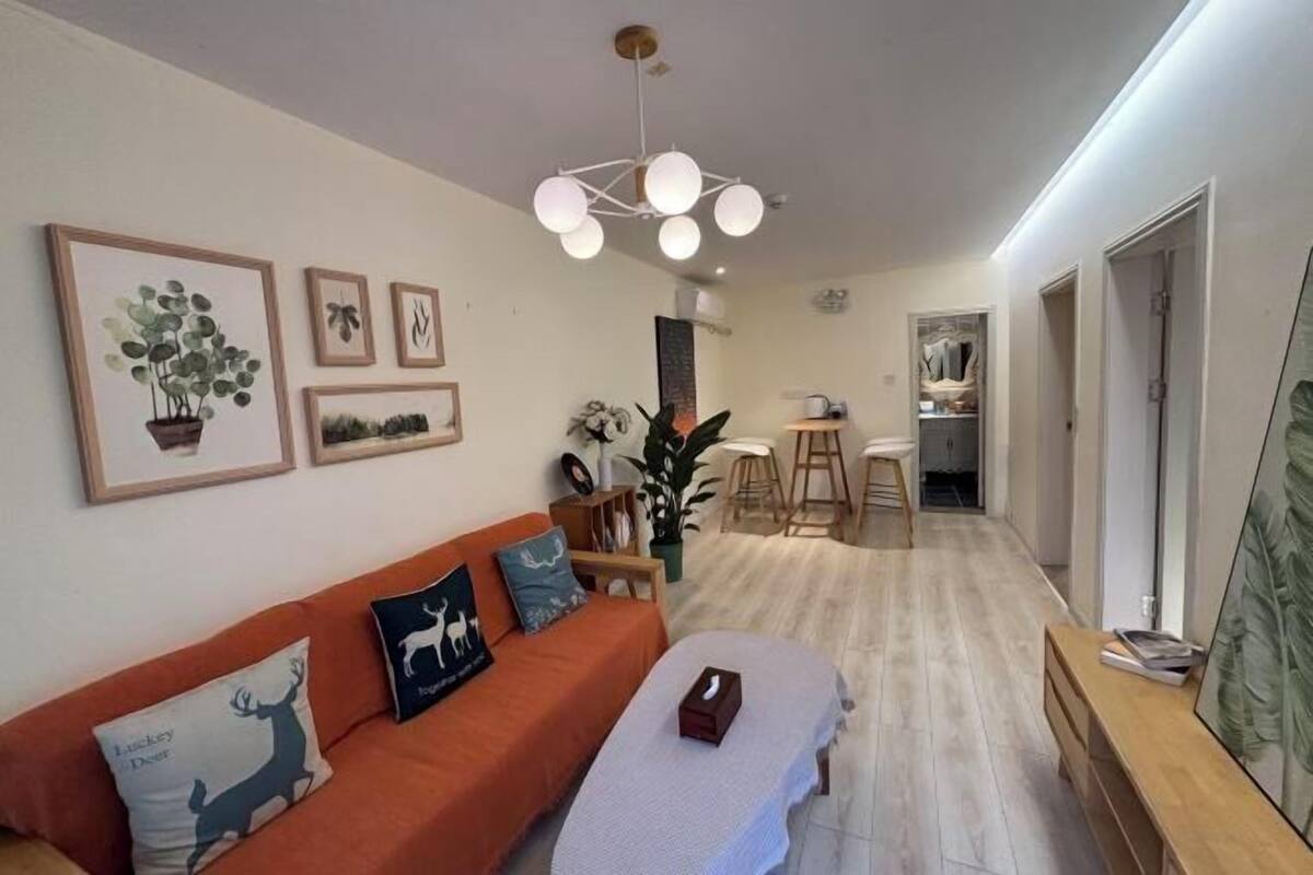 A modern living room with a sofa, a coffee table, and a dining area with stools. There are framed pictures on the wall and a ceiling light fixture.
