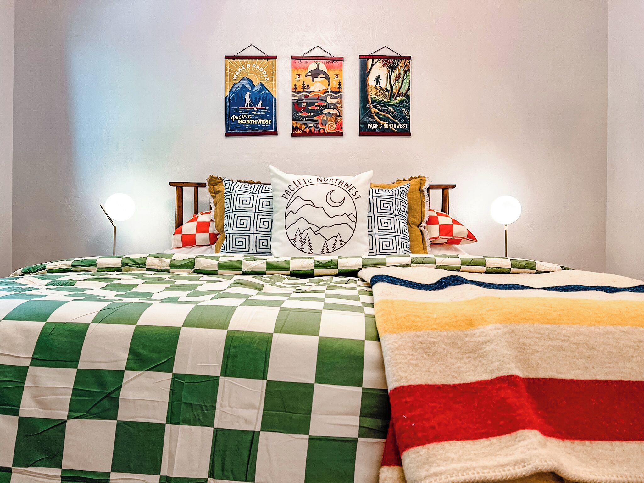 Ein ordentlich bezogenes Bett mit karierter Tagesdecke, mehreren Kissen und einer Wand mit drei Postern zum Thema Nordwest-Pazifik.