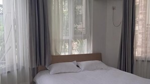 Egy hálószoba, amelyben van egy ágy, függönyök, egy ablak és egy a falra szerelt telefon.