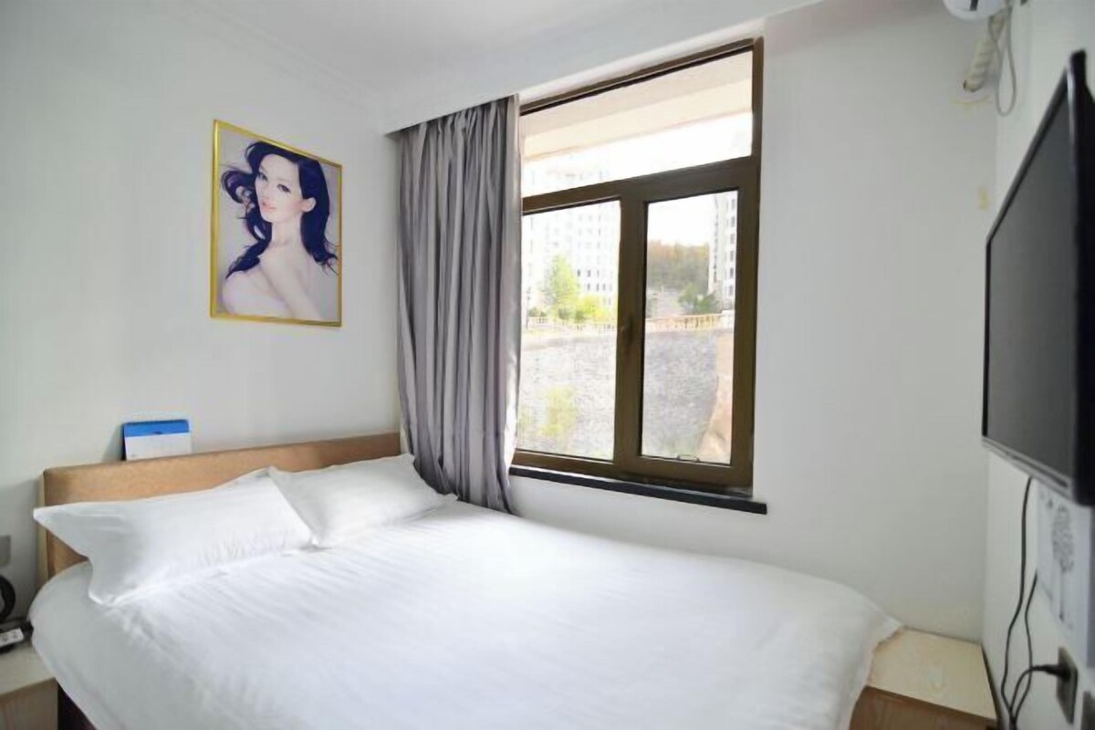 A hotel room with a bed, a window with curtains, a painting on the wall, and a television on the right side.