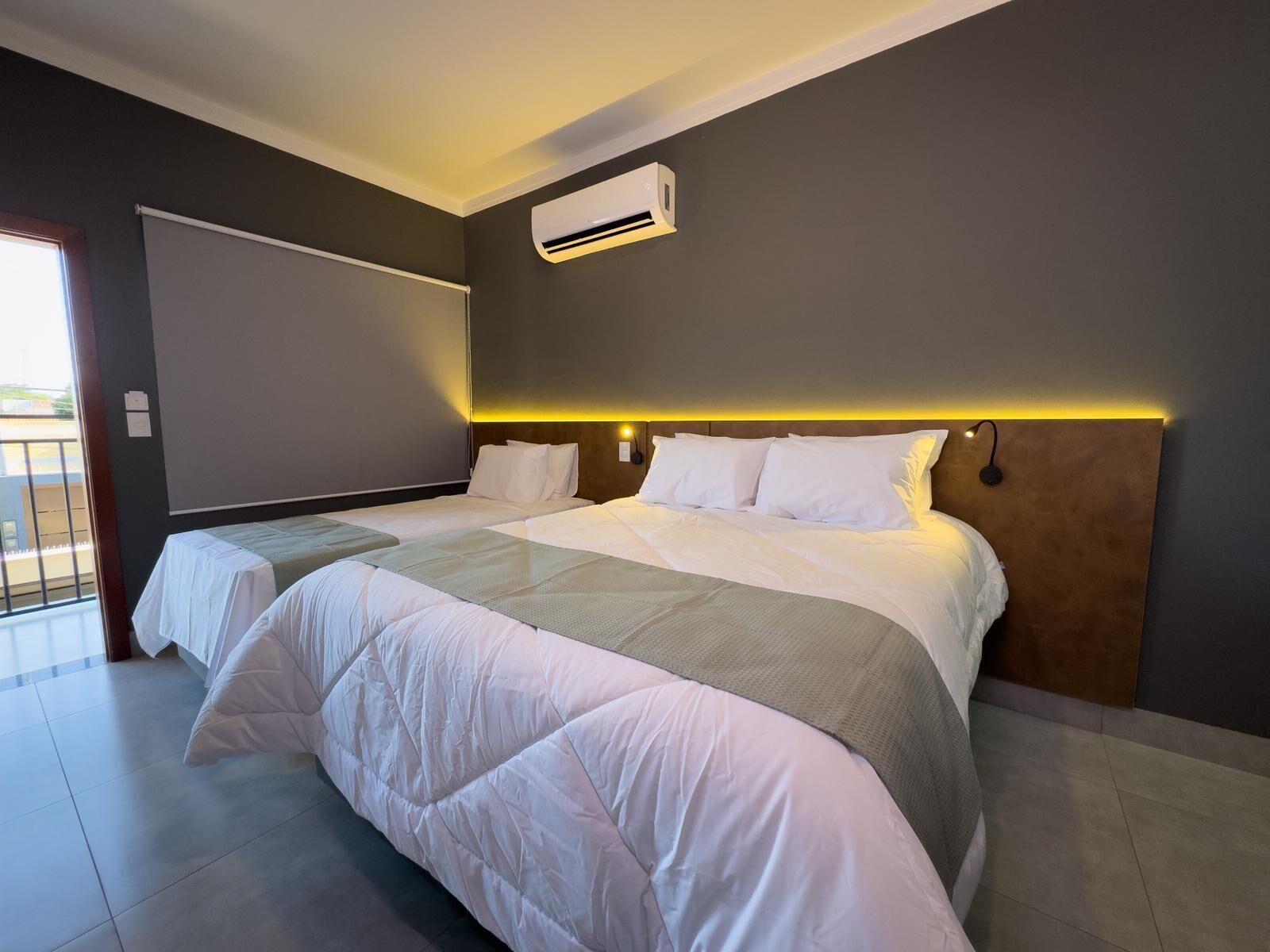 A modern hotel room with a large bed, a bedside table, an air conditioning unit, and a view through a sliding door.