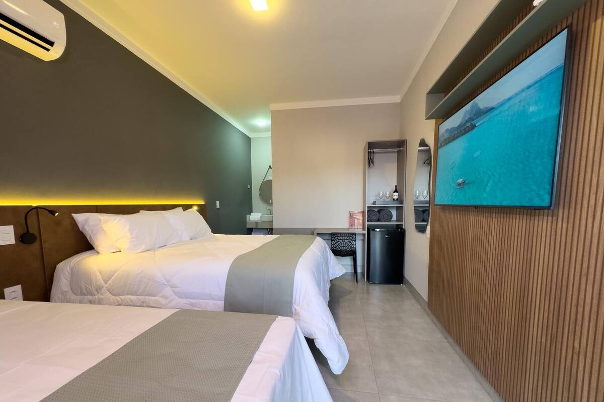 Quarto de hotel com duas camas, uma escrivaninha, um espelho, uma lixeira e um quadro na parede.