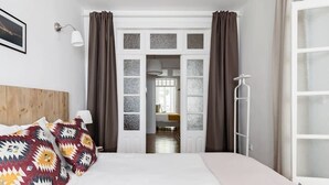 Un dormitori amb un llit, un capçal de llit, una finestra amb cortines, una làmpada penjada a la paret i un radiador.
