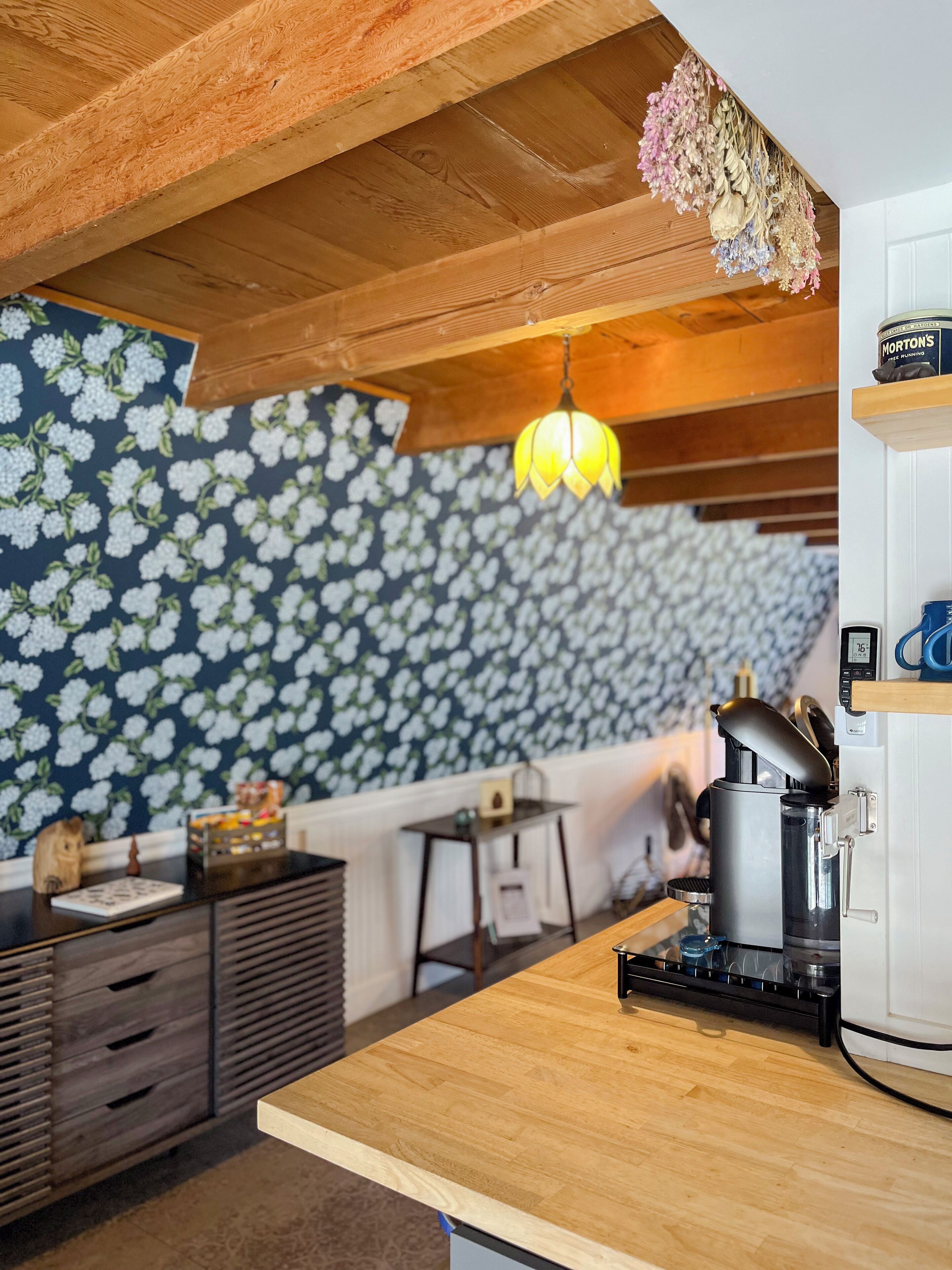 A kitchen with a wooden countertop, a coffee machine, a hanging light fixture, and a floral wallpaper.