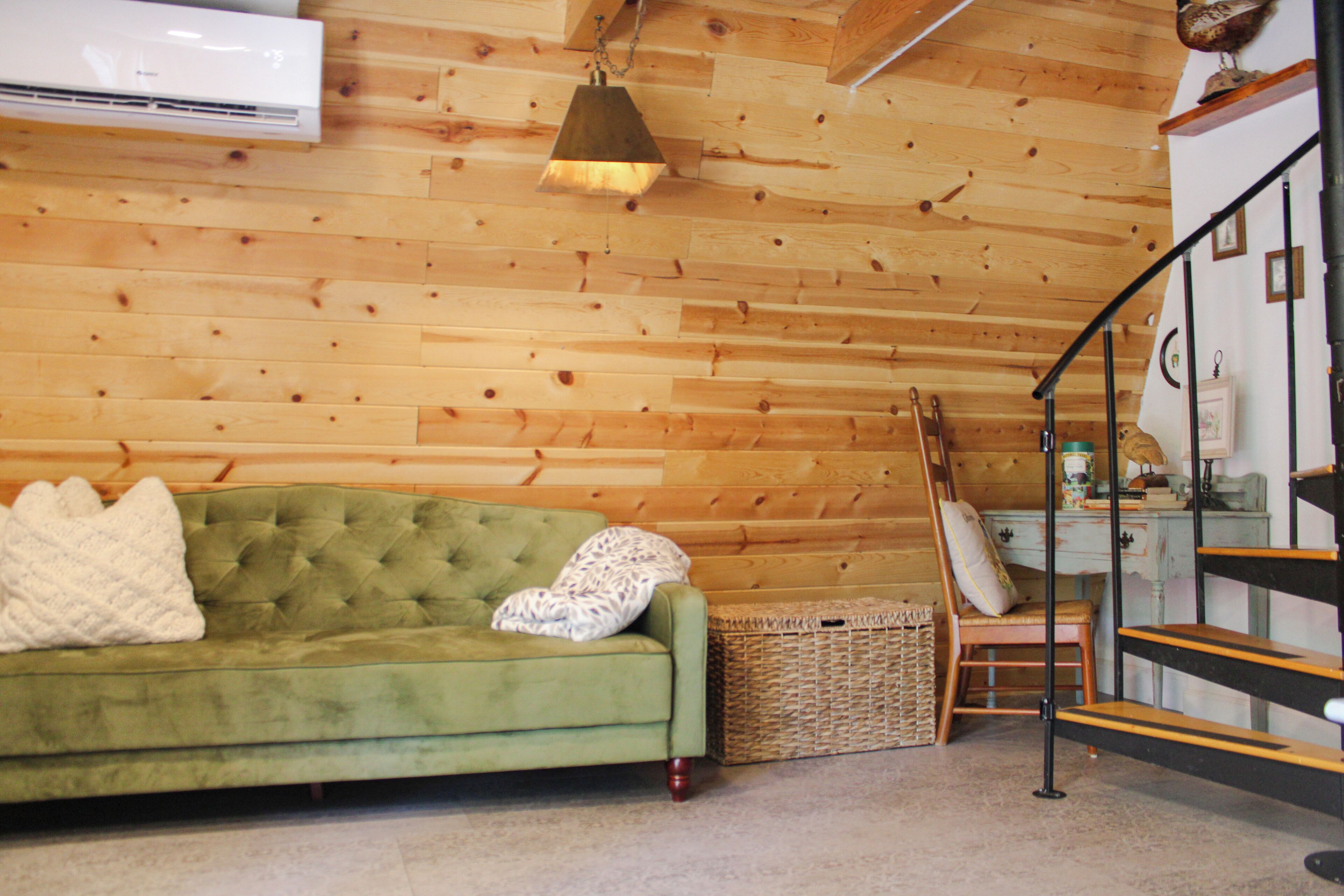 Una habitación con un sofá verde, una cesta de mimbre, una escalera de madera y un aire acondicionado de pared.