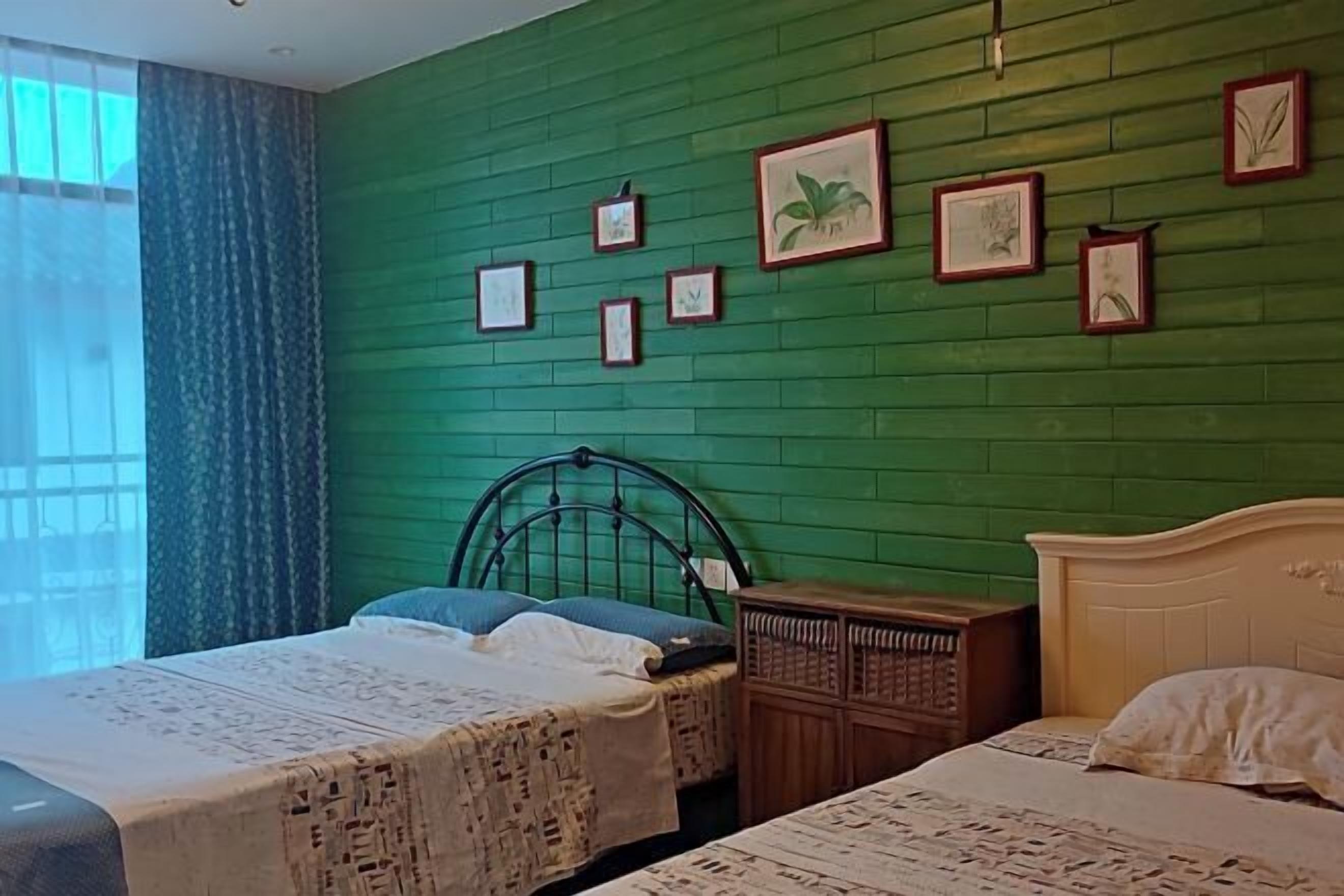 Une pièce avec un mur vert, deux lits recouverts de couvre-lits à motifs, une table de chevet en bois et des tableaux encadrés.