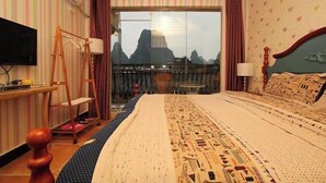 A hotel room with a bed, a television, a wooden desk, a ladder, and a view of mountains through a glass door.