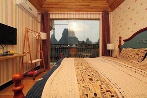 A hotel room with a bed, a television, a wooden desk, a ladder, and a view of mountains through a glass door.