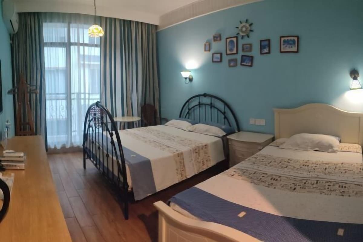 A room with two beds, a desk, a chair, a window with curtains, and wall-mounted lights.