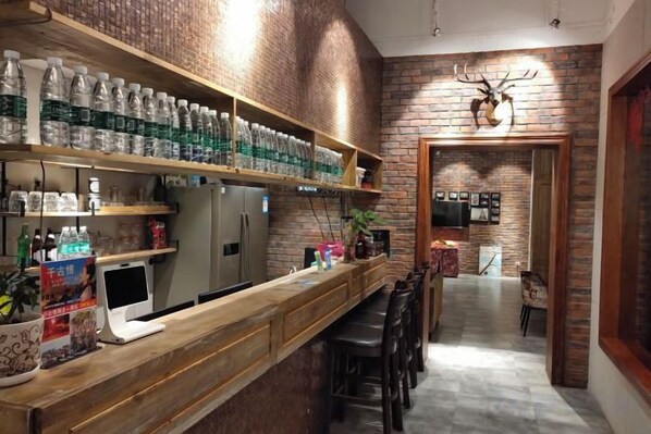 A bar area with a long counter, shelves with bottled beverages, a refrigerator, and a seating area with stools.
