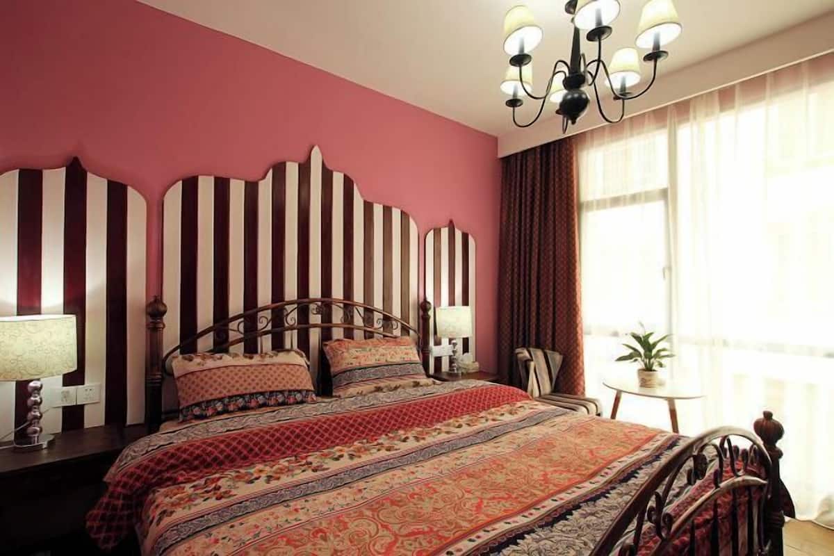 A bedroom with a striped headboard, a patterned bedspread, a chandelier, and a small table with a plant.