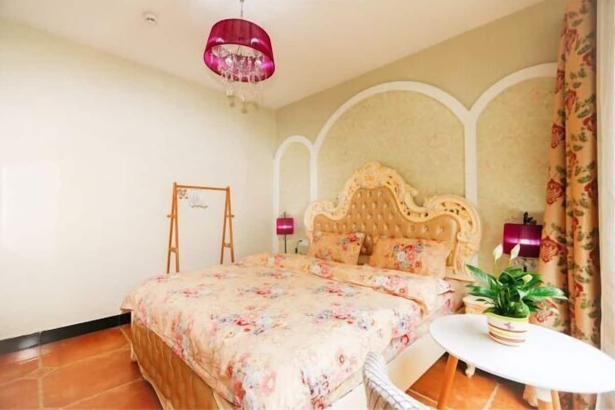 A bedroom with a floral bedspread, a wooden headboard, a small round table with a plant, and a pink chandelier.