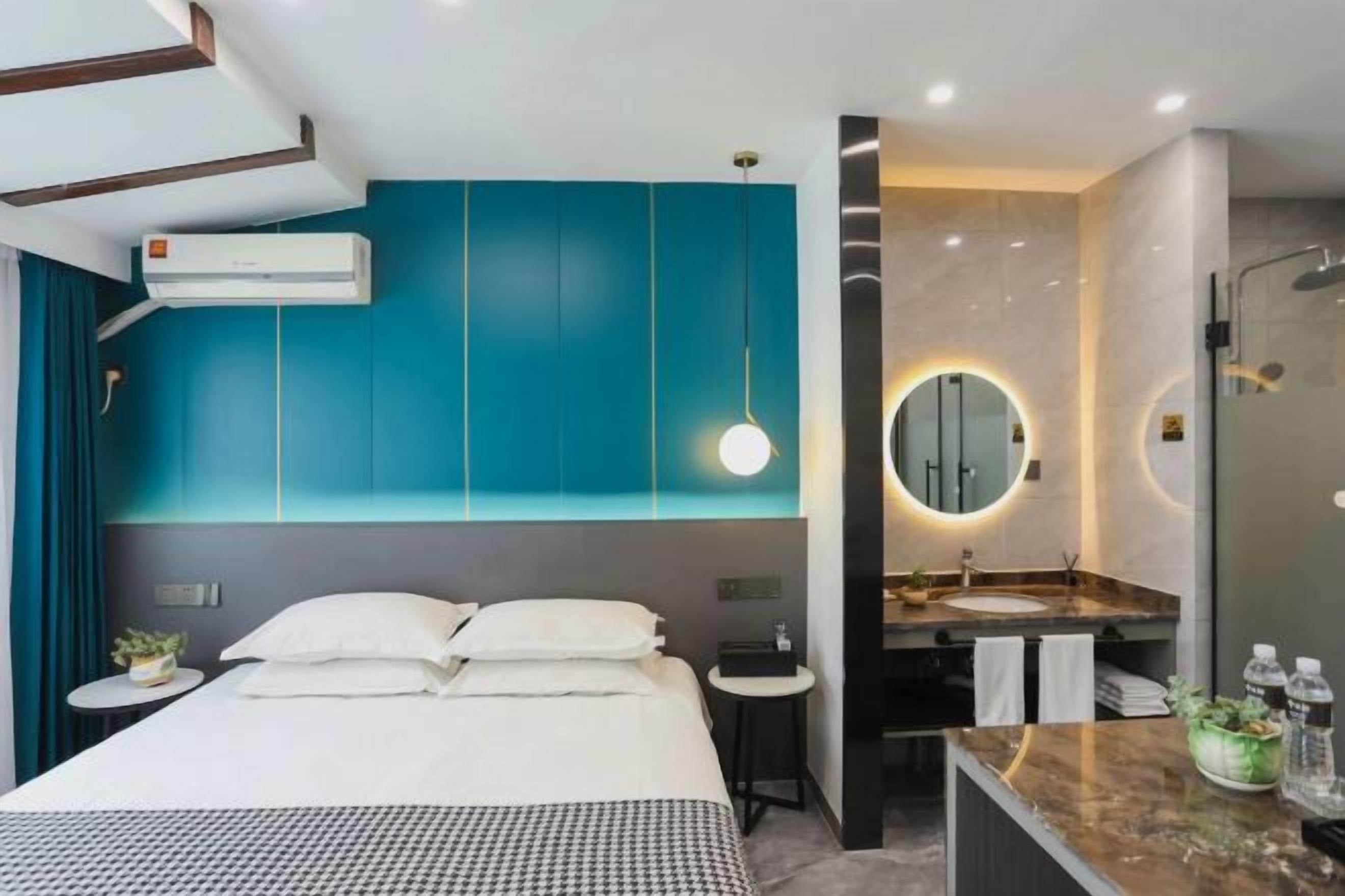 Una habitación de hotel moderna con una cama grande, un espejo redondo, un baño con lavamanos y ducha, y paredes color turquesa.