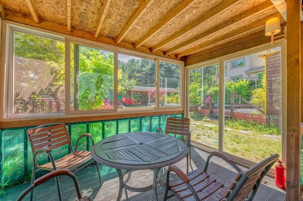 Une terrasse couverte avec une table ronde et quatre chaises, de grandes fenêtres et une vue sur un jardin et les maisons voisines.