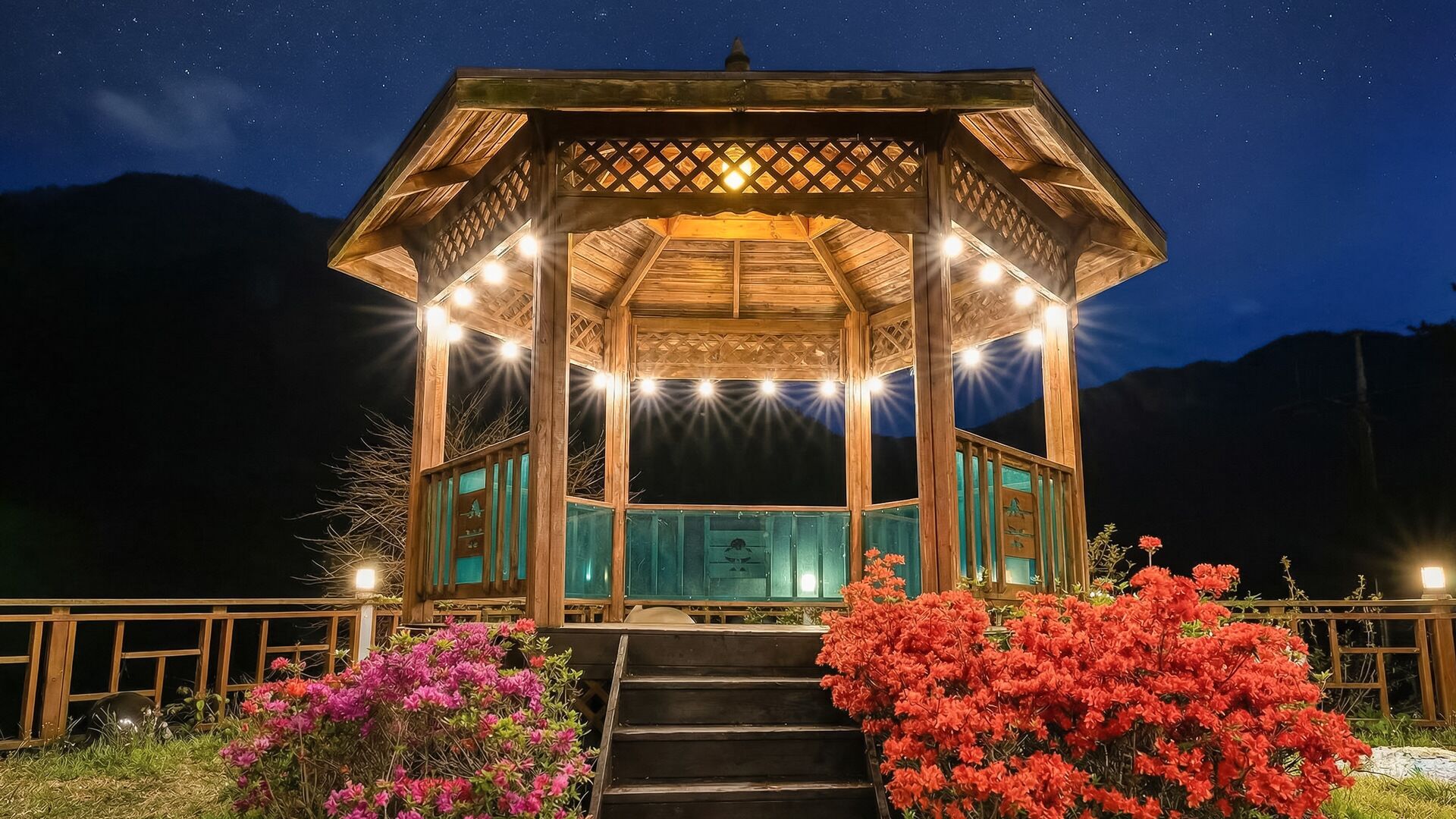 A wooden gazebo with a starry night sky in the background, surrounded by vibrant flowers and a mountain range.