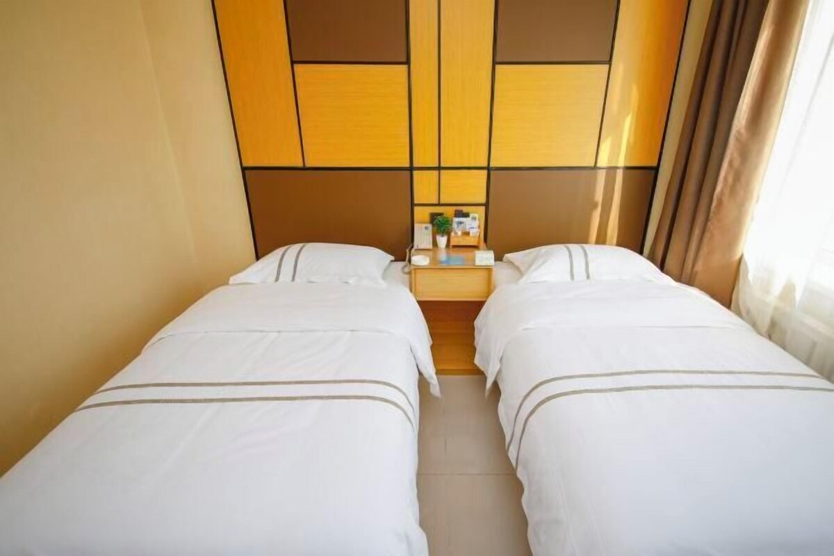 A hotel room with two single beds, a wooden headboard, a bedside table with a lamp, and a window with curtains.