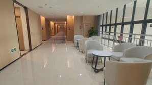Een hotelgang met een lange hal, zithoek met stoelen en tafels, en een groot raam met luxaflex.