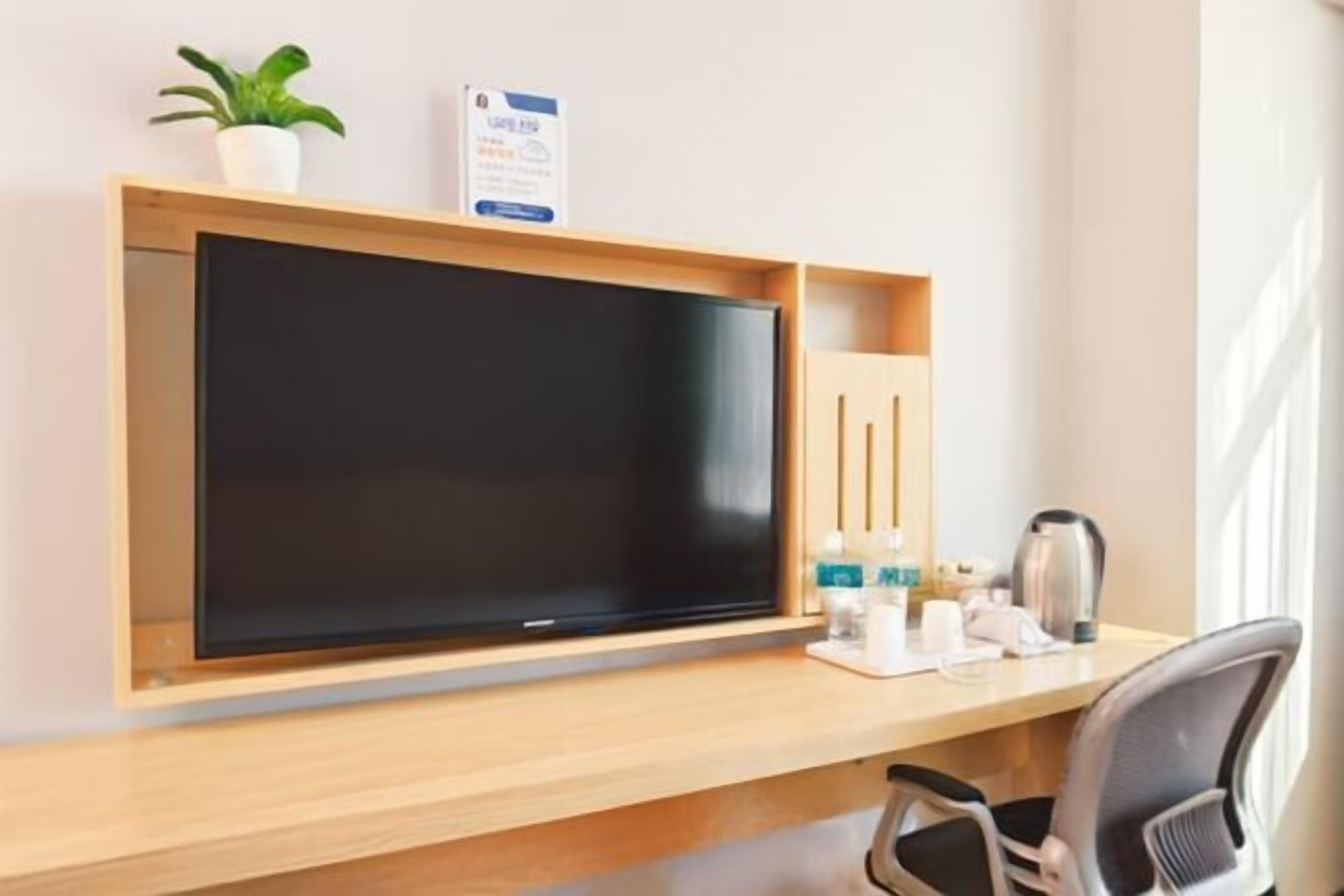 A hotel room with a wooden desk, a flat-screen TV, a small potted plant, a sign, a kettle, and a chair.