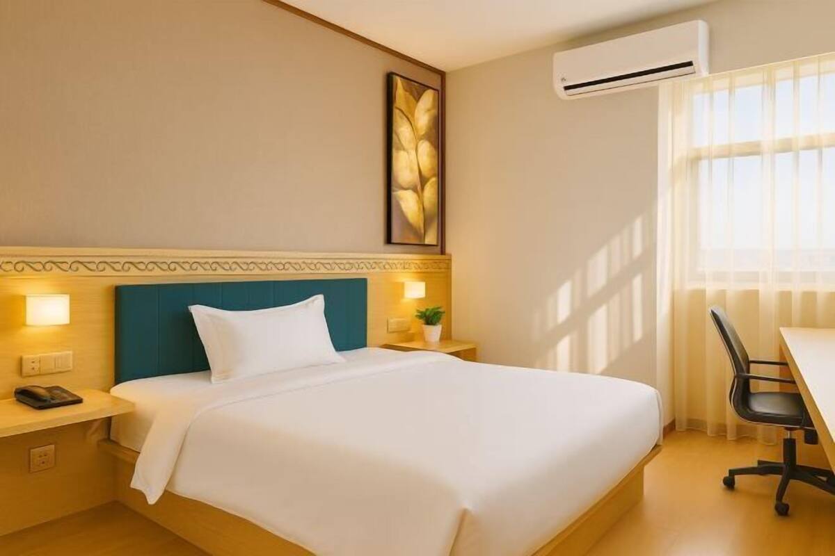 A hotel room with a bed, bedside table, lamp, air conditioning unit, and a window with curtains.