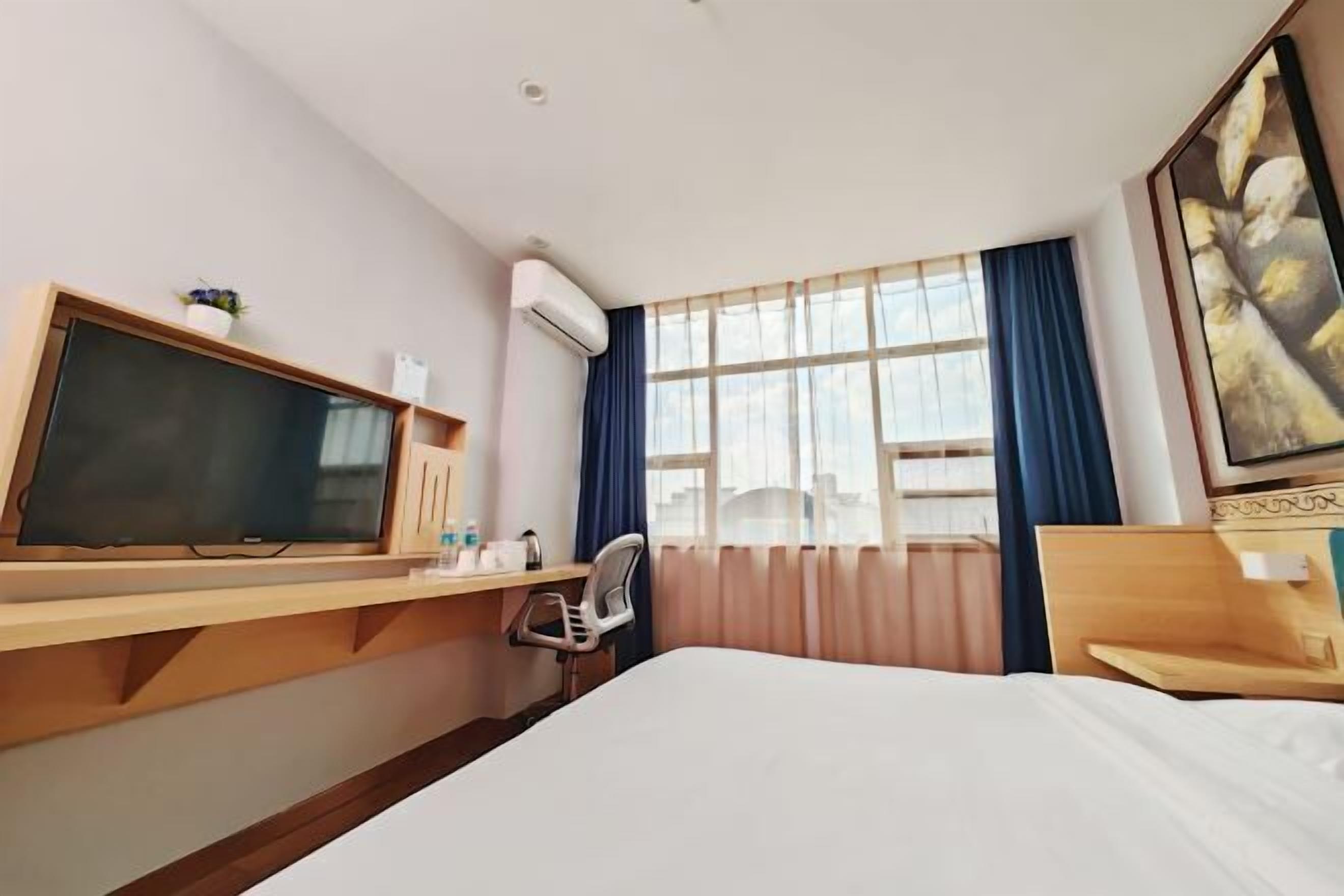 A hotel room with a bed, a desk with a TV, a chair, a window with curtains, and an air conditioning unit.