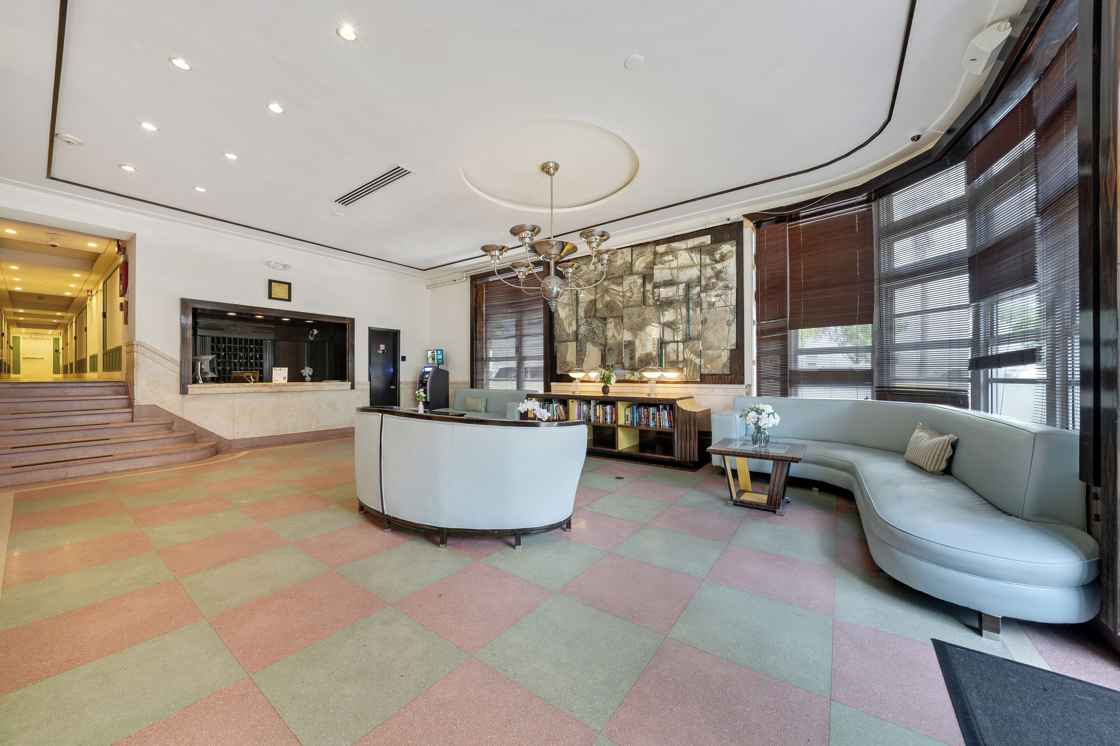 A modern lobby with a reception desk, a curved sofa, a chandelier, and a staircase leading to another area.