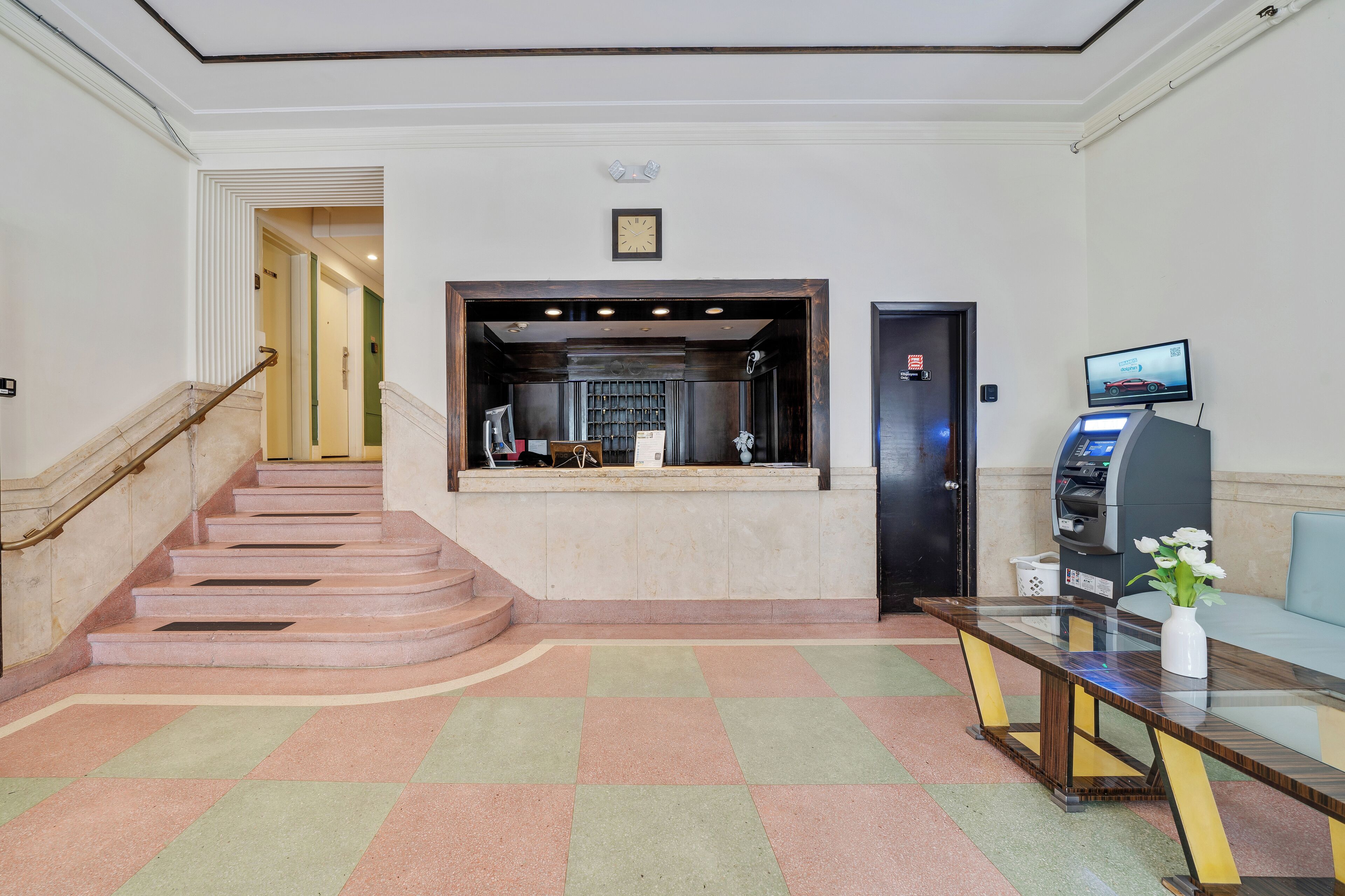 A lobby with a staircase, a reception desk, a clock, and a self-service kiosk.