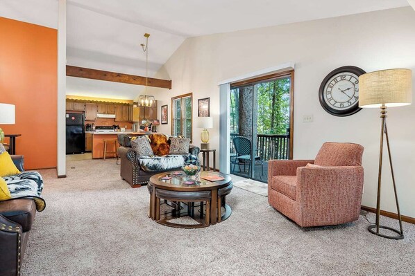 A living room with a sofa, armchair, coffee table, and a lamp. There is a kitchen in the background and a balcony with a view of trees.