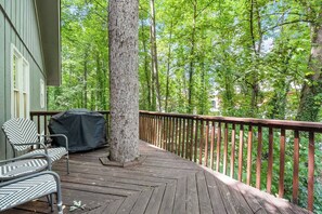 A wooden deck with a covered grill and two chairs, surrounded by a wooden railing and dense green foliage.
