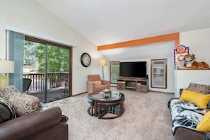 A living room with a sofa, armchair, coffee table, TV, and a balcony with a view.