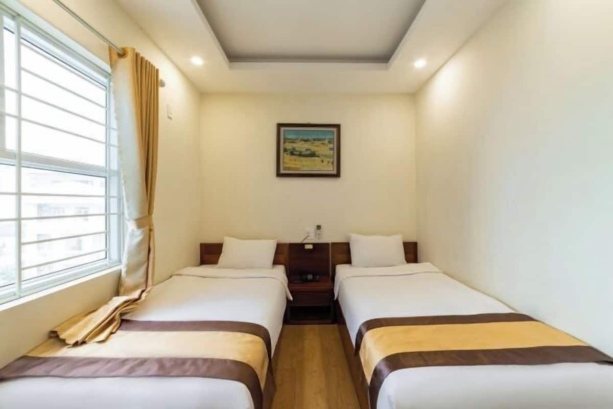 Une chambre avec deux lits, une table de chevet, un tableau au mur et une fenêtre avec des rideaux.
