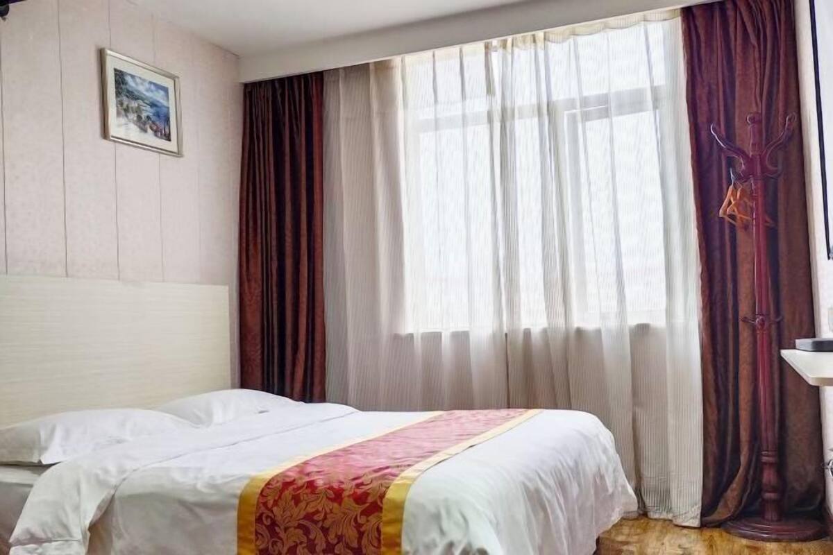 Hotellihuone, jossa on sänky, ikkuna verhoilla, naulakko ja taulu seinällä.