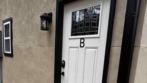 Una puerta blanca con la letra 'B' encima, una pequeña ventana con diseño de rejilla, y un felpudo con la palabra 'WELCOME' en el suelo.