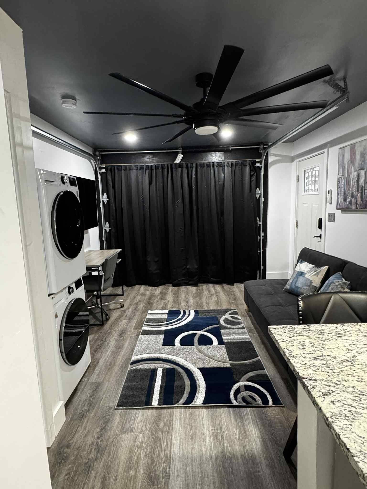 A compact living space with a ceiling fan, a washing machine, a dark curtain, a patterned rug, a sofa, and a granite countertop.