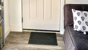 Una puerta blanca con un ventanal pequeño, un felpudo negro y un sofá de cuero marrón con un cojín estampado.