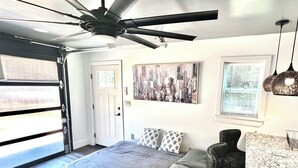 Un dormitorio con una cama, un ventilador de techo, una pintura de paisaje urbano, una silla y una ventana con persianas.