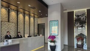 Egy modern szállodai recepció, amely tartalmaz egy recepciós pultot, két alkalmazottat, díszítő falpaneleket, egy képkeretbe helyezett műalkotást és egy virágtálban elhelyezett növényt.