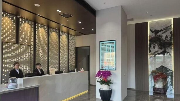 En modern hotellobby med en receptionsdisk, två anställda, dekorativa väggpaneler, ett inramat konstverk och en krukväxt.