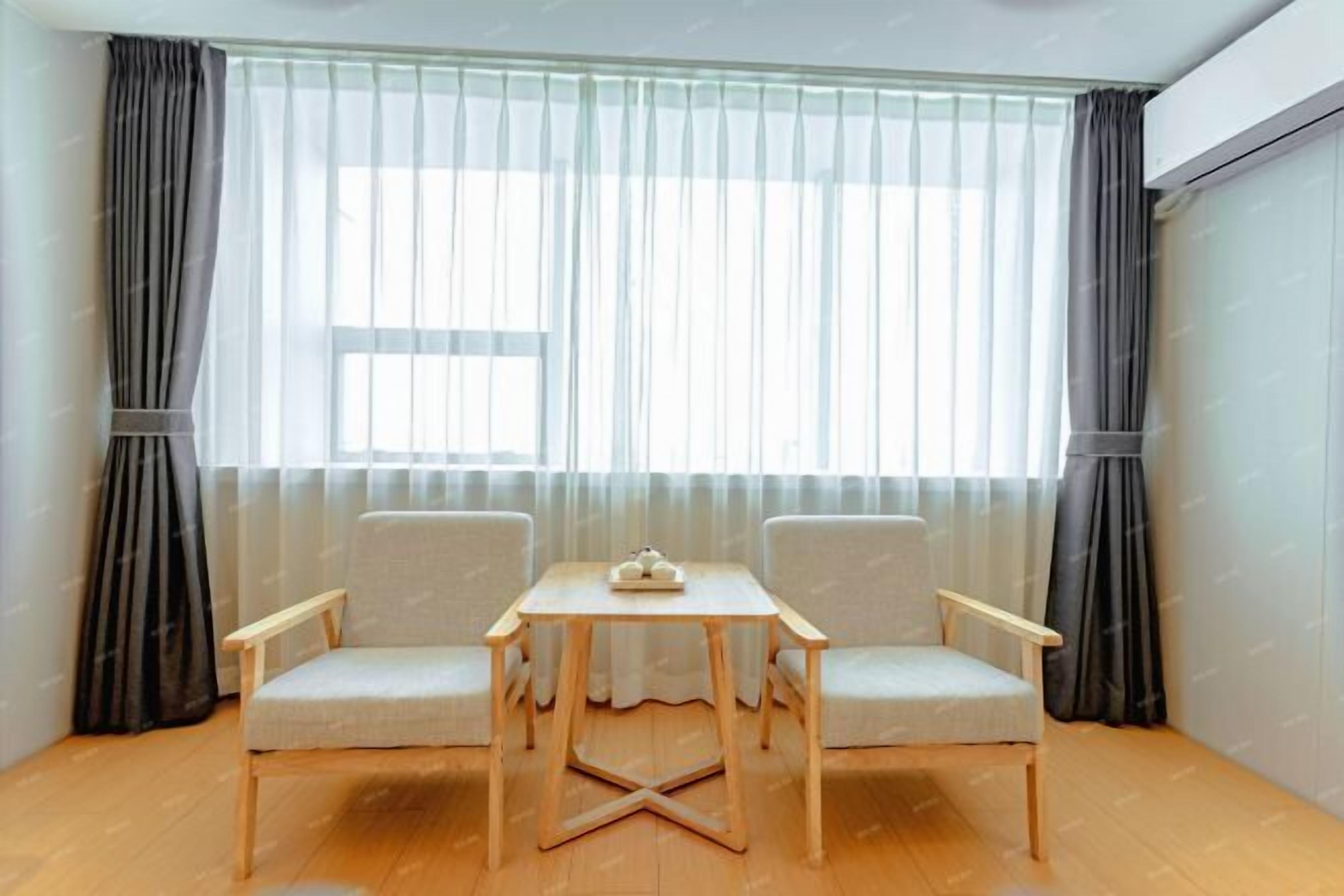 Una habitación con piso de madera, dos sillas, una mesita y un ventanal con cortinas translúcidas.