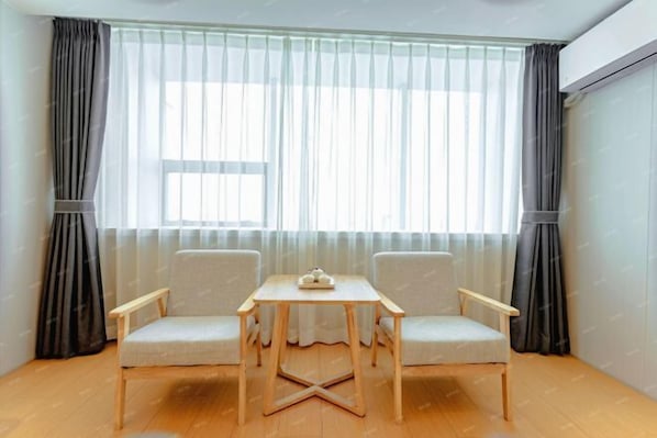 Ein Zimmer mit Holzboden, zwei Stühlen, einem kleinen Tisch und einem Fenster mit durchsichtigen Vorhängen.