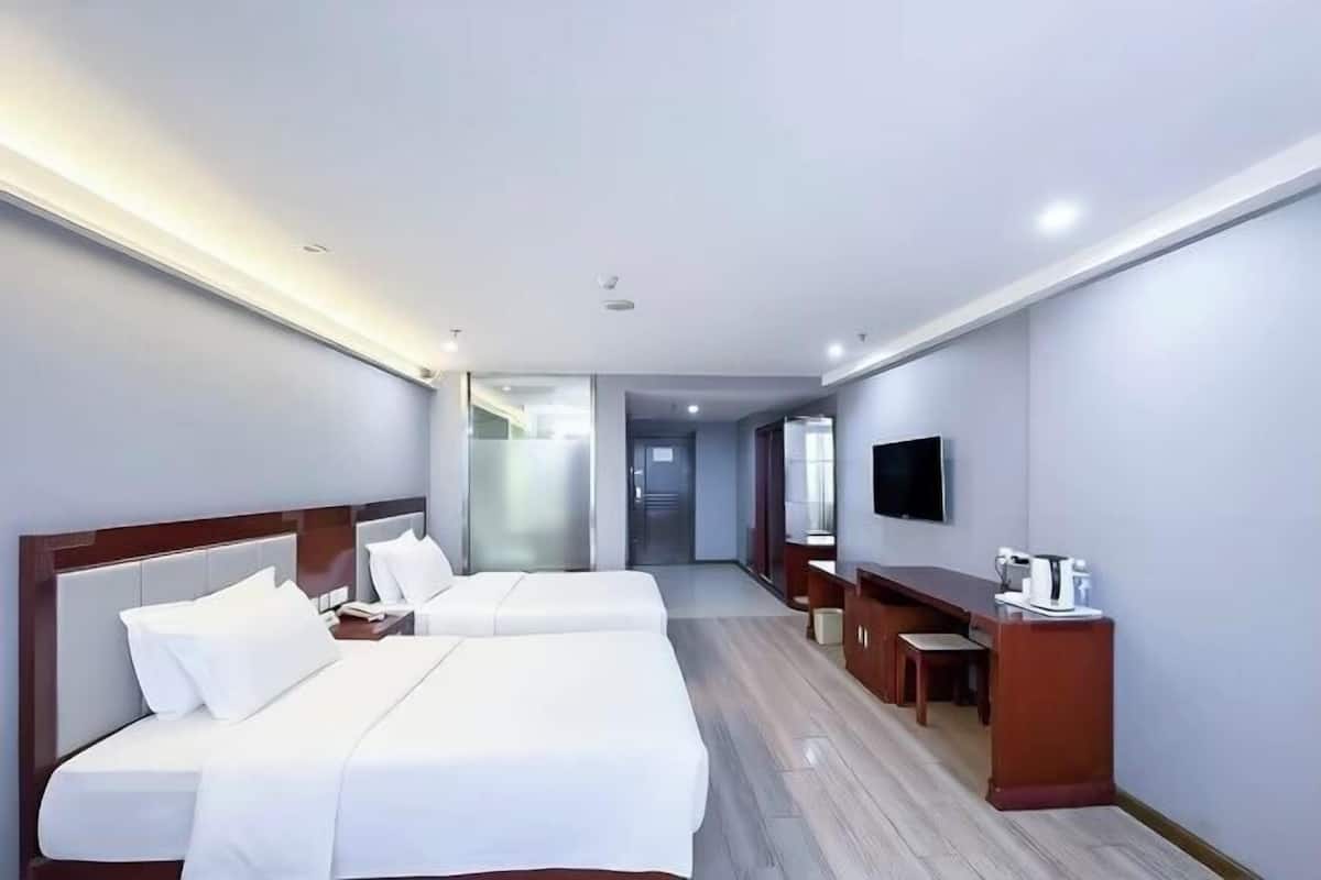 A hotel room with two beds, a desk, a television, and a bathroom visible through a glass door.