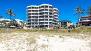 A multi-story hotel with balconies, located near a beach with sand dunes and palm trees.