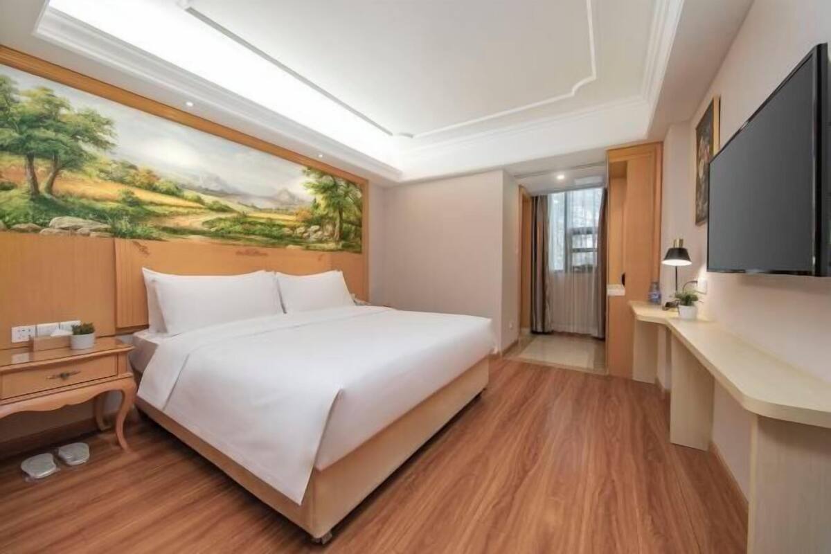 A hotel room with a bed, wooden flooring, a painting on the wall, a desk, and a TV.