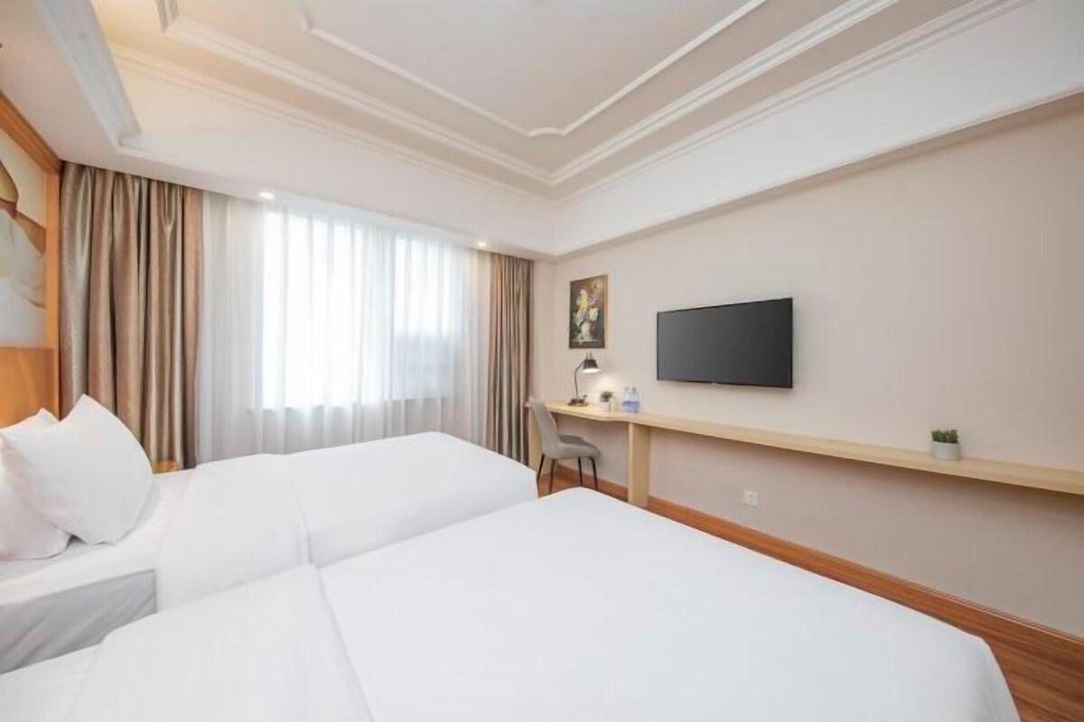 A hotel room with two beds, a desk, a flat-screen TV, and a window with curtains.